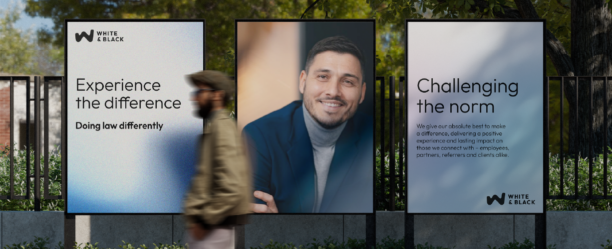 Three outdoor posters on a fence, featuring White & Black branding and messages including “Experience the difference” and “Challenging the norm,” with a person walking past in the foreground.