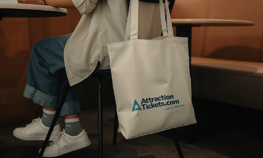 A person sitting in a café holds a canvas tote bag printed with the AttractionTickets.com logo.