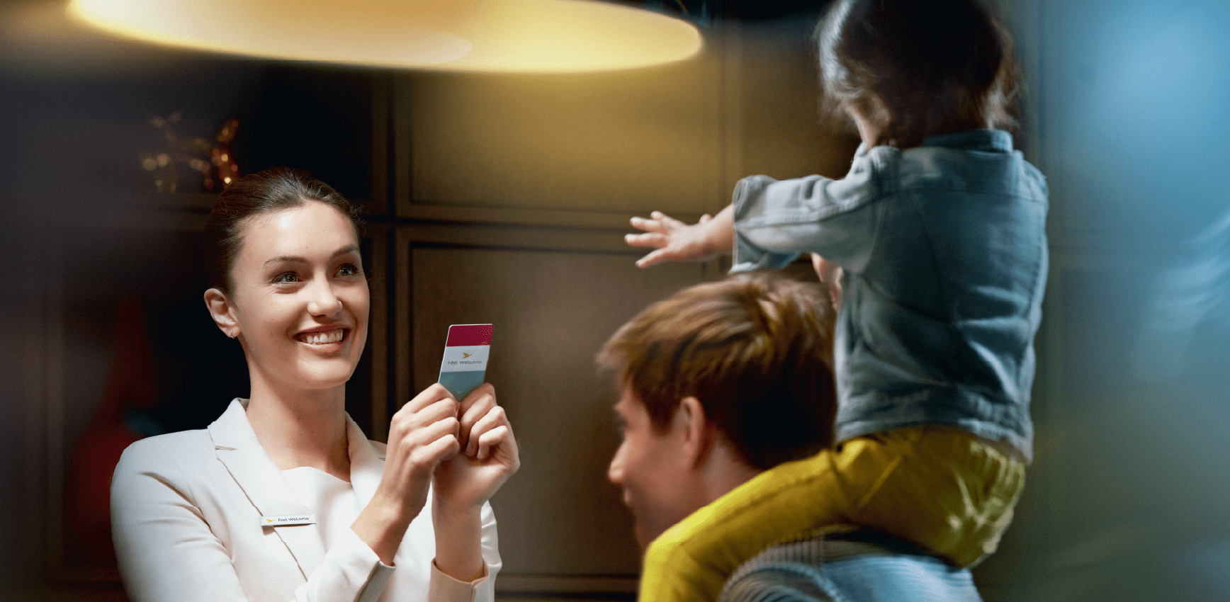 A hotel staff member shows a room key card to an adult carrying a child on their shoulders.
