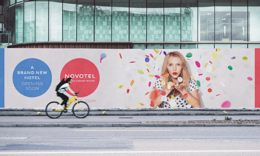"A brand new Novotel hotel opening soon” hoarding featuring a woman blowing colourful confetti, with a cyclist passing by in front.