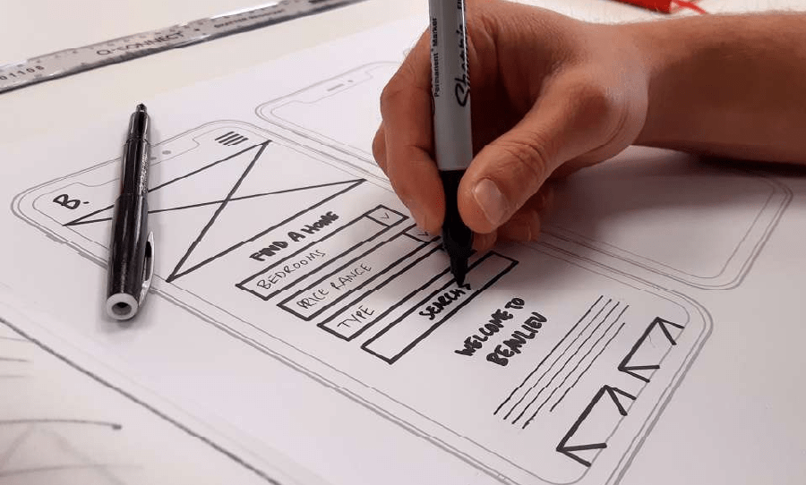 A hand draws a mobile app wireframe with a marker, sketching search fields and layout elements on paper.