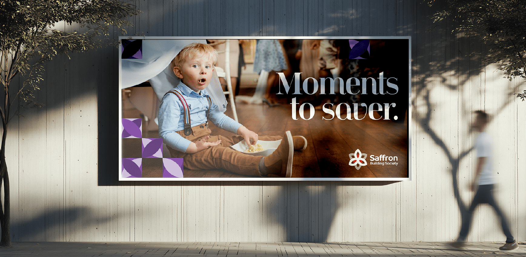 A Saffron Building Society billboard showing a child sitting on a wooden floor at an event, with the headline “Moments to saver.”