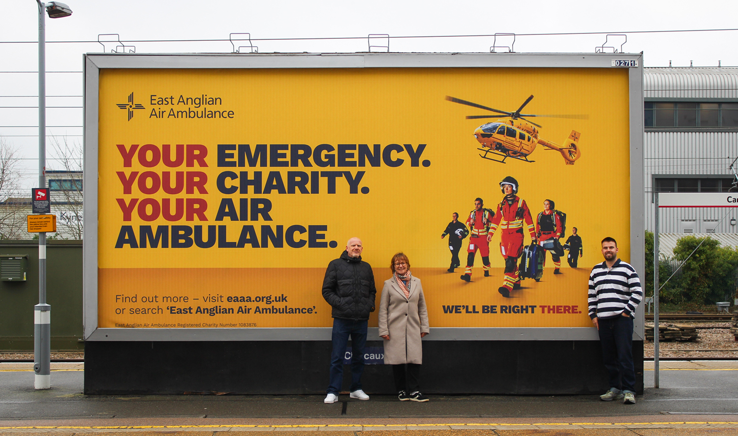Large yellow East Anglian Air Ambulance billboard at a train station featuring bold text and rescue crew imagery, with three people standing in front of it.