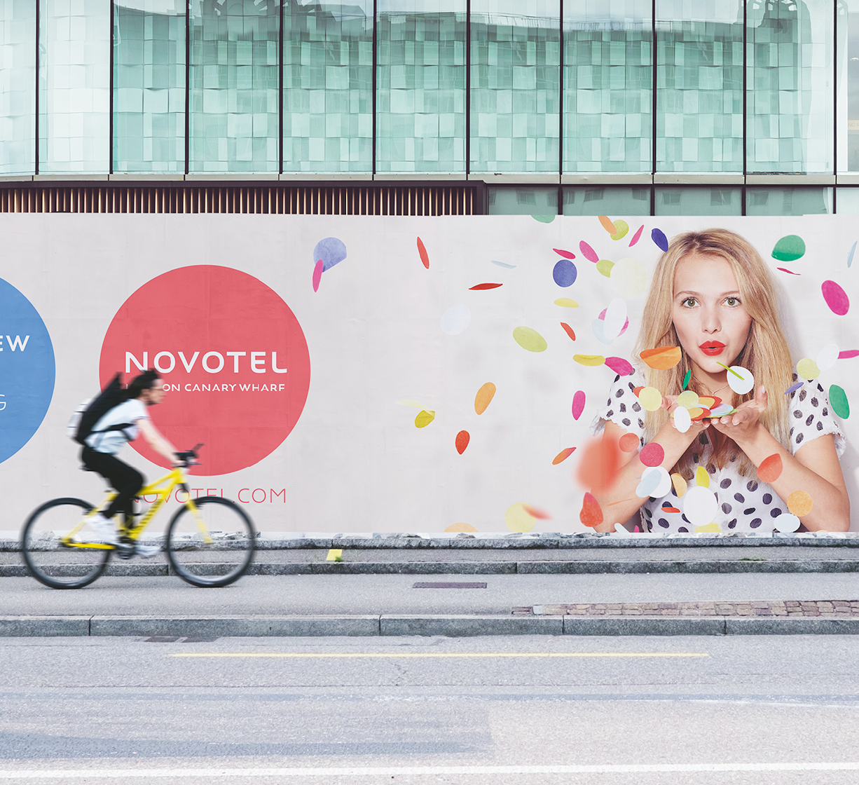 Large Novotel Canary Wharf advertisement on a building façade with colorful confetti graphics, while a cyclist rides past on the street.