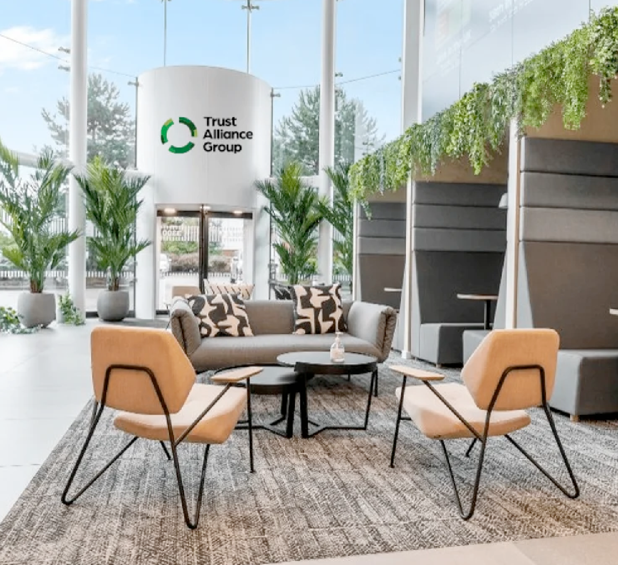 Modern lobby area with light chairs, a sofa, indoor plants, and a wall displaying the Trust Alliance Group logo.