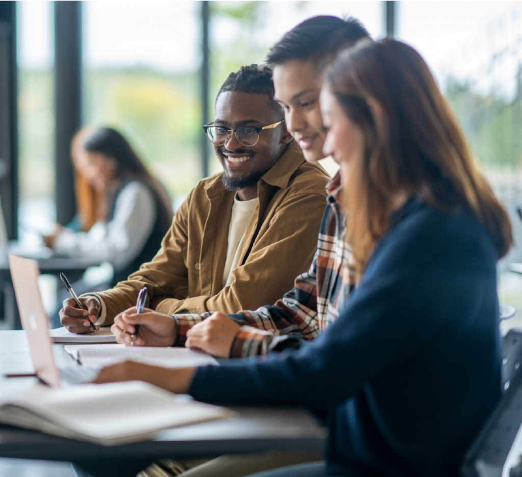 Group of students sitting at desks in a classroom, writing in notebooks and using a laptop.