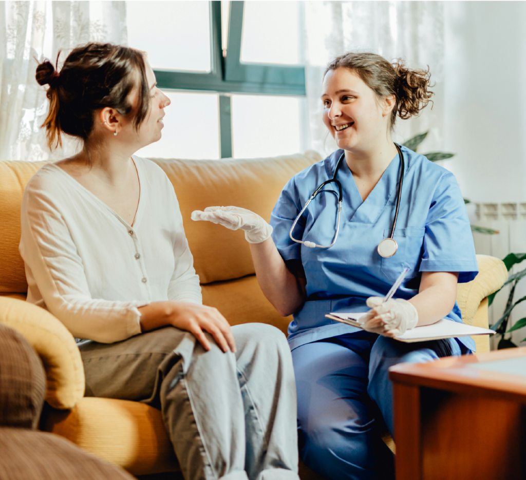 A healthcare professional in scrubs speaks with a person seated on a sofa while holding a clipboard.