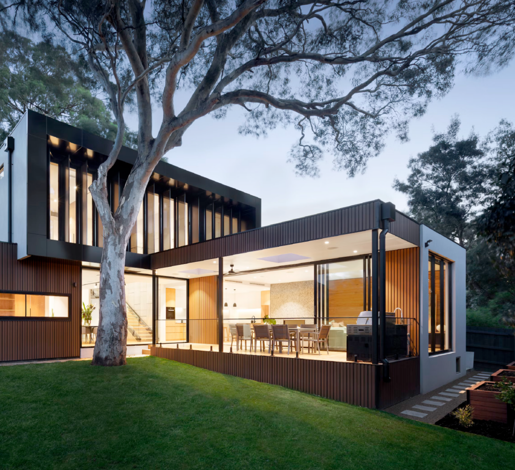 Modern two‑storey house with large glass walls, wood paneling, and an open indoor–outdoor living area beside a tall tree.