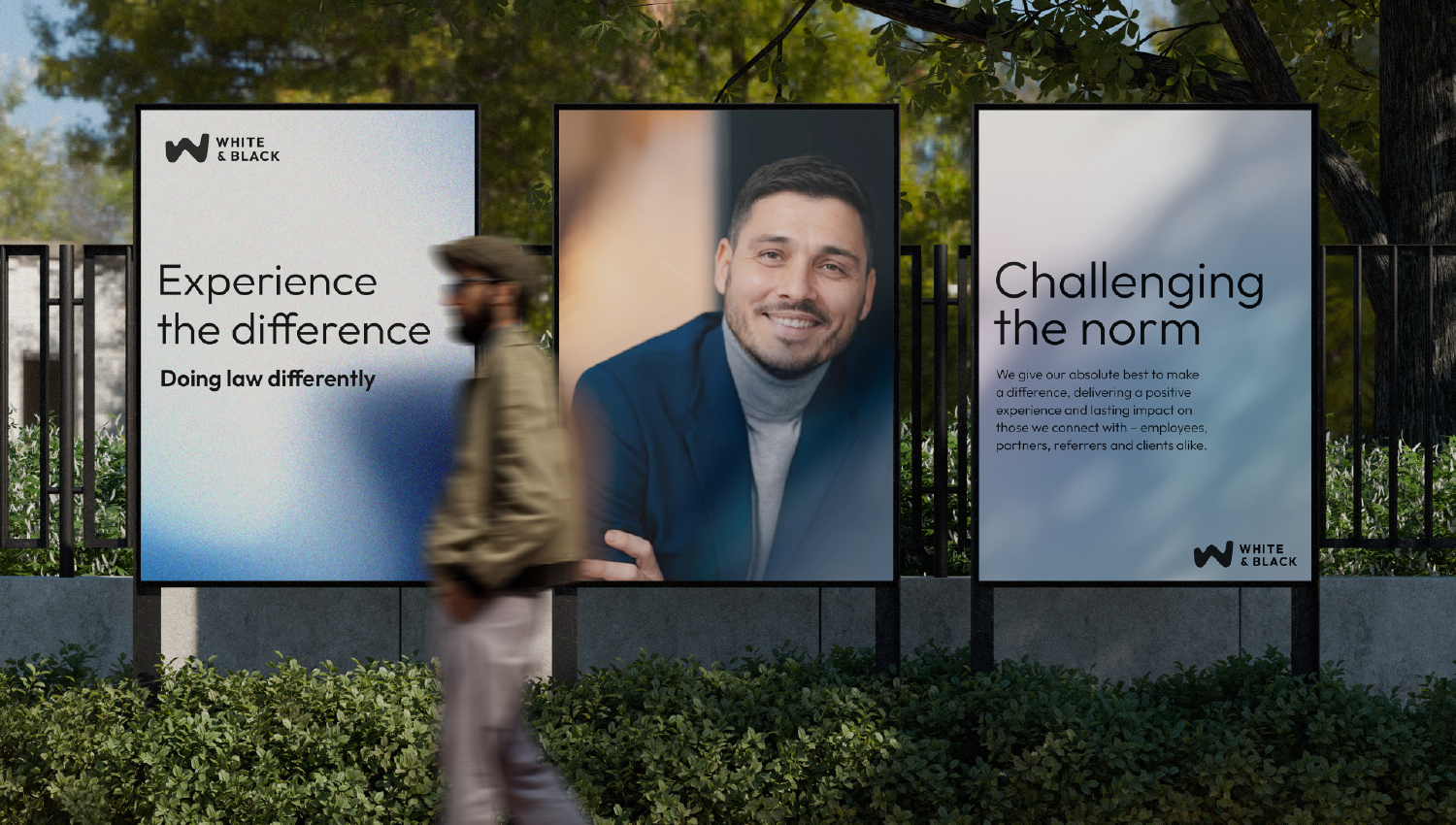 Three outdoor posters on a fence, featuring White & Black branding and messages including “Experience the difference” and “Challenging the norm,” with a person walking past in the foreground.