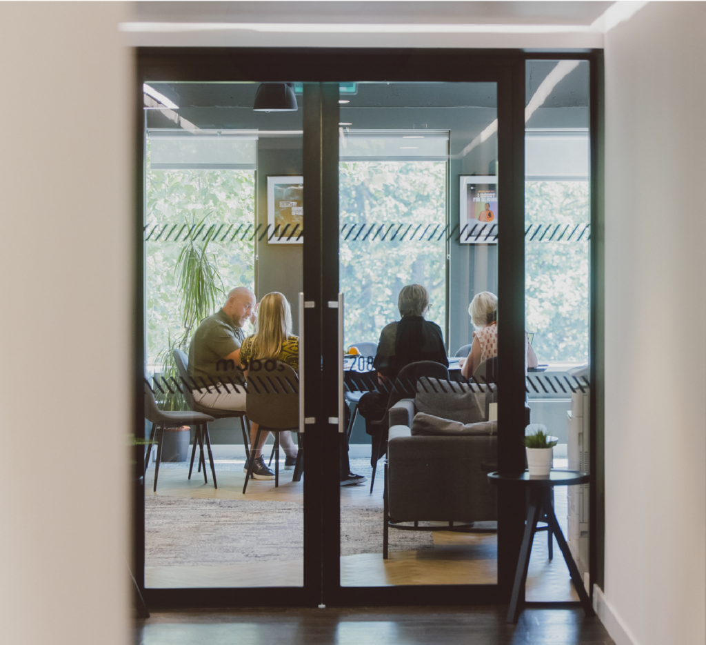 View through glass doors of a small meeting room where several people are seated and talking around a table.