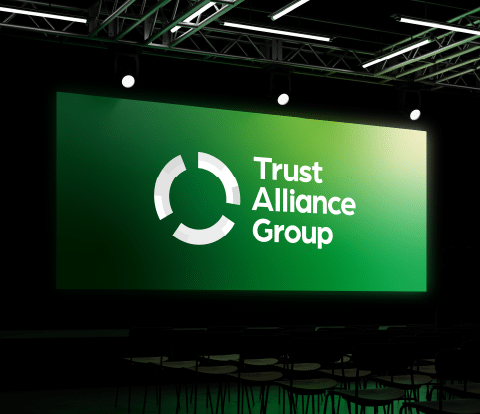 Trust Alliance Group logo displayed on a large green illuminated screen in a dark event space with seating in the foreground.