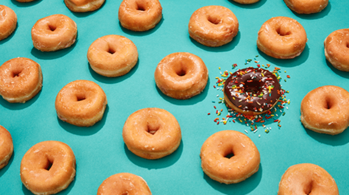 A collection of plain glazed doughnuts arranged in a pattern on a bright turquoise background, with one chocolate-iced doughnut covered in colourful sprinkles standing out among them.